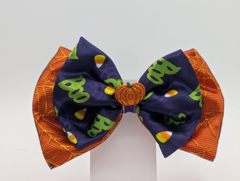Puede incluir: Un lazo para el pelo con tem&aacute;tica de Halloween con un centro azul marino con patrones de "Boo" y caramelos de ma&iacute;z. Tiene detalles laterales naranjas y un adorno de calabaza brillante. Un accesorio festivo para Halloween.