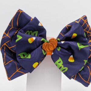 Puede incluir: Un lazo para el pelo con tem&aacute;tica de Halloween. La tela azul marino presenta telara&ntilde;as naranjas y un estampado de caramelos de ma&iacute;z y la palabra "Boo" en verde. El lazo tiene un centro de calabaza con purpurina naranja.