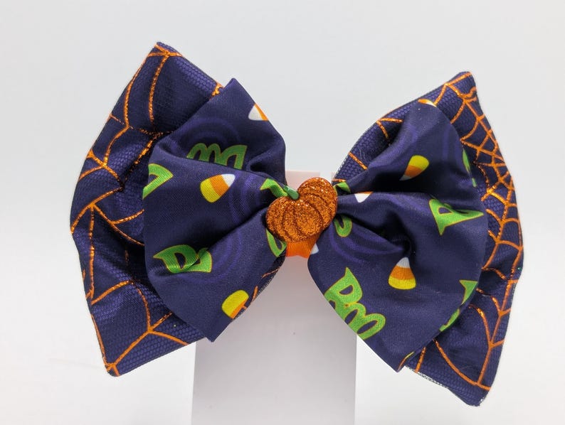 Puede incluir: Un lazo para el pelo con tem&aacute;tica de Halloween. La tela azul marino presenta telara&ntilde;as naranjas y un estampado de caramelos de ma&iacute;z y la palabra "Boo" en verde. El lazo tiene un centro de calabaza con purpurina naranja.