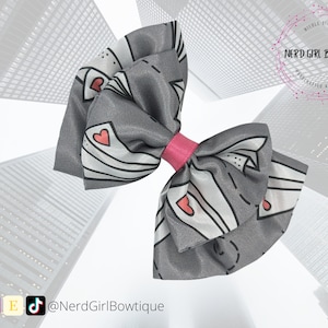 May include: A gray and white bow with a pink center, featuring a pattern of stylized envelopes with hearts. The bow is against a blurred background of city buildings. The text "Nerd Girl Bowtique" is visible in a circle at the top of the image.