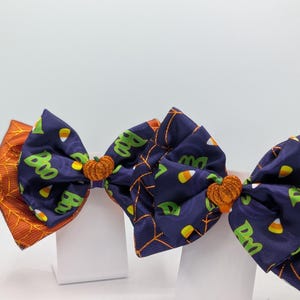 Puede incluir: Lazos para el pelo con tem&aacute;tica de Halloween. Los lazos presentan una tela azul marino con texto verde "Boo" y caramelos de ma&iacute;z, detalles de calabaza naranja y patrones de telara&ntilde;as y hojas naranjas. Sobre un fondo blanco.