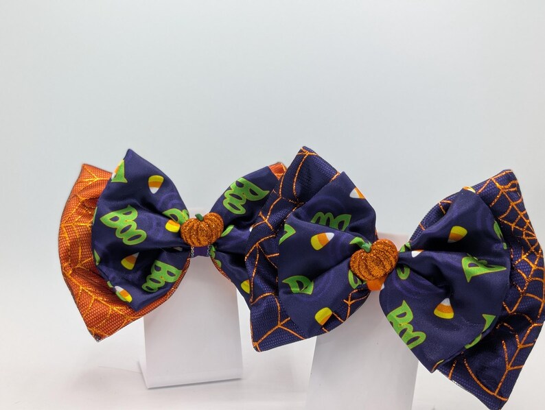 Puede incluir: Lazos para el pelo con tem&aacute;tica de Halloween. Los lazos presentan una tela azul marino con texto verde "Boo" y caramelos de ma&iacute;z, detalles de calabaza naranja y patrones de telara&ntilde;as y hojas naranjas. Sobre un fondo blanco.