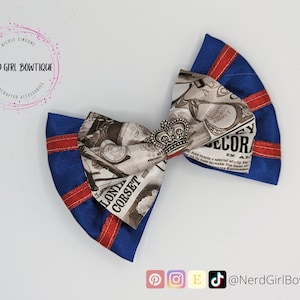 May include: A blue and red bow with a newspaper print design and a silver crown charm. The bow is handmade and features the text "LOND CORSET" and "DECOR" in the newspaper print.