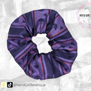 May include: Purple and red patterned scrunchie with a geometric design. The scrunchie is made of satin fabric and has a  "Nerd Girl Bowtique" logo.