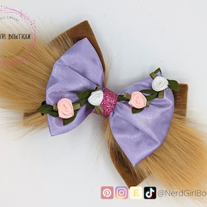 Puede incluir: Un lazo para el cabello hecho a mano con un lazo de satén morado claro con detalles de flores rosas y blancas. El lazo tiene una base de fieltro marrón y está adornado con extensiones de cabello sintético largas de color marrón claro. De Nerd Girl Bowtique.