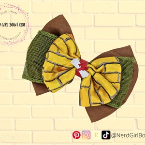 Puede incluir: Un lazo grande y en capas para el cabello con un centro estampado amarillo y gris, acentos de arpillera verde y fieltro marrón. Un adorno blanco y rojo está en el centro. El lazo está hecho a mano.