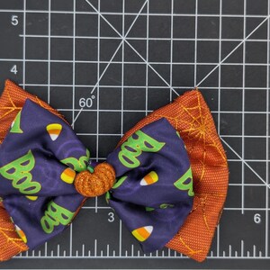 Puede incluir: Un lazo tem&aacute;tico de Halloween con un centro en forma de calabaza. El lazo presenta un centro azul oscuro con patrones de "Boo" y caramelos de ma&iacute;z, superpuesto a una tela naranja con detalles de telara&ntilde;a. El lazo mide unos 10 cm de ancho.