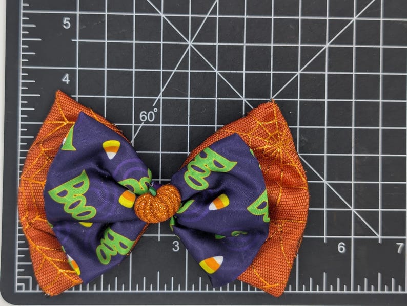 Puede incluir: Un lazo tem&aacute;tico de Halloween con un centro en forma de calabaza. El lazo presenta un centro azul oscuro con patrones de "Boo" y caramelos de ma&iacute;z, superpuesto a una tela naranja con detalles de telara&ntilde;a. El lazo mide unos 10 cm de ancho.