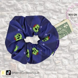 May include: A blue scrunchie featuring a repeating pattern of green skull and crossbones. A one-dollar bill is tucked into a zippered pocket on the scrunchie. The scrunchie is a handmade accessory.