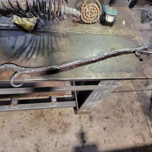 Puede incluir: Herramienta de metal forjada a mano con un mango retorcido y un gancho curvado. La herramienta de metal plateado descansa sobre un banco de trabajo de metal. La herramienta es probablemente para herrería o trabajo de metales.