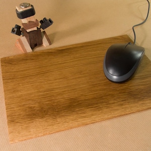 Könnte beinhalten: Ein hölzernes Mauspad mit einer schwarzen Computermaus und einer kleinen Holzroboterfigur. Das Mauspad ist rechteckig und aus hellbraunem Holz gefertigt. Die Maus ist schwarz mit grauem Rand. Der Roboter besteht aus verschiedenen Holzstücken.
