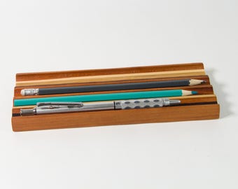 May include: A wooden pencil and pen holder with a striped design. The holder has three slots for pens and pencils. The holder is made of light and dark wood.