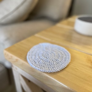 Op de afbeelding: Een lichtgrijze gehaakte onderzetter op een houten tafel.