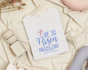 Könnte beinhalten: Weißes T-Shirt mit einer blauen und rosa Grafik, auf der "He is Risen Hallelujah" mit einem Kreuz und floralen Akzenten steht.