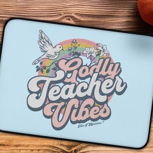 Può includere: Una custodia per laptop azzurra con bordo nero. La custodia presenta un design con una colomba, un arcobaleno e fiori, insieme al testo "Godly Teacher Vibes" in un font retrò. Il design è nei toni del bianco, rosa e blu.