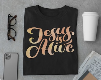 Könnte beinhalten: Schwarzes T-Shirt mit einem Aquarell-Design, auf dem in Gelb-Orange "Jesus lebt" steht.