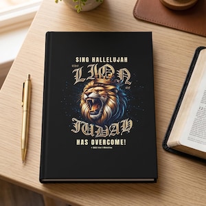May include: A black hardback journal featuring a graphic of a roaring lion wearing a crown, with the text "SING HALLELUJAH THE LION OF JUDAH HAS OVERCOME!" in gold lettering. The journal is positioned next to a gold pen and an open book.