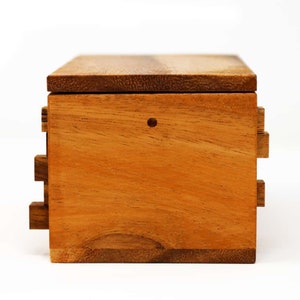 May include: A wooden puzzle box with a rectangular shape. The box is made of light brown wood with a darker brown lid. The sides of the box have a series of interlocking pieces. A small hole is visible on the front of the box.