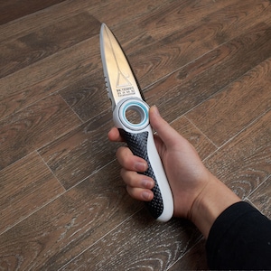 Può includere: Un coltello nero e argento con un anello blu attorno al manico. Il coltello ha una lama seghettata e un manico nero con un'impugnatura bianca.