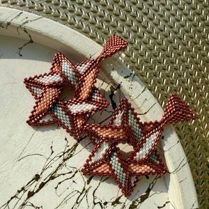 May include: Handmade beaded star earrings in shades of red, white, and peach. The earrings are made with small beads and have a geometric design. The earrings are displayed on a white tray with black paint splatters.