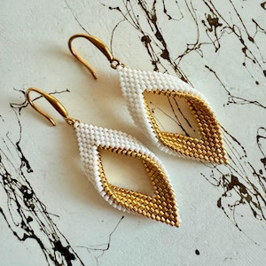 May include: A pair of handmade earrings with a leaf-like design. The earrings feature a white beaded exterior and a gold beaded interior, with gold-colored ear wires. The earrings are displayed on a white surface with black paint splatters.