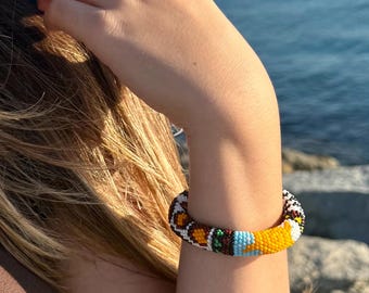 May include: A beaded bracelet with a vibrant pattern of orange, yellow, brown, white, green, and blue beads. The bracelet is worn on a person's wrist, with the ocean in the background. The bracelet is a colorful accessory.