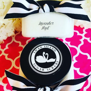 May include: A black jar with a white label that says "Swan Island Sugar Scrubs" and a white swan logo. The jar is tied with a black and white striped ribbon. The jar is filled with a white sugar scrub. The label also says "Lavender Mint".