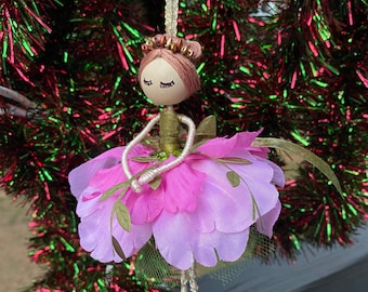 May include: A handmade Christmas ornament in the shape of a ballerina. The doll has a wooden head with closed eyes, a pink and green floral skirt, and gold accents. The ornament hangs from a gold cord against a backdrop of a Christmas tree.