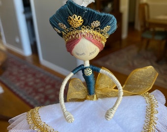 Peut inclure: Ornement de Noël fait à la main en forme de ballerine. La poupée a une tête en bois, une couronne bleu canard et dorée, et un tutu blanc avec une bordure dorée. Les bras sont en fil de fer crème. Un nœud doré est à la taille. Ruban argenté pour suspendre.