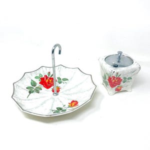 May include: A white, two-tiered serving tray with a silver handle and a matching sugar bowl. The tray and bowl are decorated with red rose floral patterns and green leaves. The tray has a silver trim.