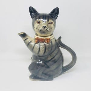 May include: A ceramic cat-shaped teapot with a grey and black striped body, cream-coloured face, and a small orange bow tie. The teapot has a spout, handle, and lid, set against a white background.