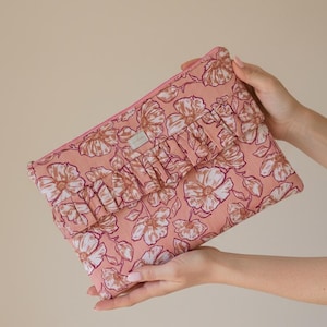 Può includere: Una pochette rosa floreale con dettaglio a balze. La pochette presenta un motivo ripetuto di grandi fiori nei toni del bianco e del marrone. La cerniera è rosa e la pochette è tenuta da due mani.