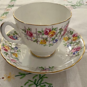 May include: A white teacup and saucer with a floral pattern of pink, yellow, and blue roses and green leaves on a white lattice background. The edges of the cup and saucer are trimmed with gold.