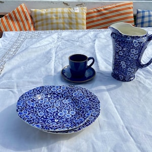 May include: A blue and white floral patterned pitcher and teacup with saucer on a white tablecloth. The teacup and saucer are on a matching blue and white floral patterned plate.