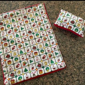 Pode incluir: Um conjunto temático de Natal com um tapete retangular e uma pequena almofada. O tecido branco apresenta um padrão repetido de ícones festivos, como árvores de Natal e presentes, delineados em preto. As bordas são aparadas em vermelho.