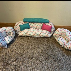 May include: A miniature sofa and two armchairs set for pets. The furniture features a white fabric with a colorful chevron pattern. The sofa has two teal and pink pillows, and a teal bolster. The chairs have the same chevron pattern.