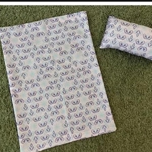 Pode incluir: Um conjunto de cobertor e almofada para bebé com um padrão azul claro e branco. O padrão apresenta formas de folhas azuis repetidas. O cobertor retangular e a almofada são apresentados numa superfície verde.