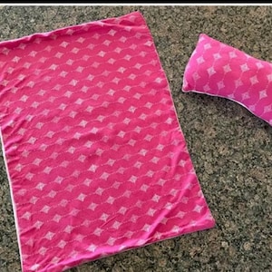 Pode incluir: Um conjunto de cobertor e almofada rosa estampados. O cobertor é retangular e a almofada tem formato retangular. Ambos os itens apresentam um padrão de diamante branco repetido em um fundo rosa.