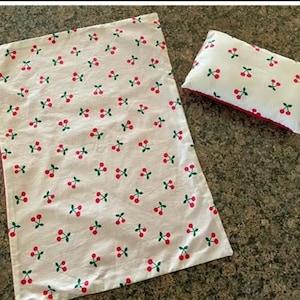 Pode incluir: Tecido retangular branco e uma pequena almofada com um padrão de cerejas vermelhas. O tecido tem uma borda vermelha. As cerejas são vermelhas com caules verdes, espalhadas sobre o fundo branco.