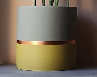 May include: A two-tone cylindrical planter. The top half is light grey, the bottom half is yellow, and a copper band separates the colours. Green plants are visible inside the planter.