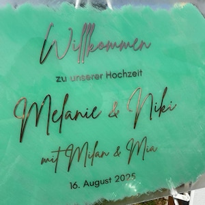 May include: A clear acrylic wedding sign with the words "Willkommen" and "Melanie & Niki" in a decorative font. The background is a light green color with the date "16. August 2025" at the bottom.
