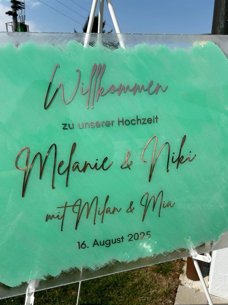 May include: A clear acrylic wedding sign with the words "Willkommen" and "Melanie & Niki" in a decorative font. The background is a light green color with the date "16. August 2025" at the bottom.