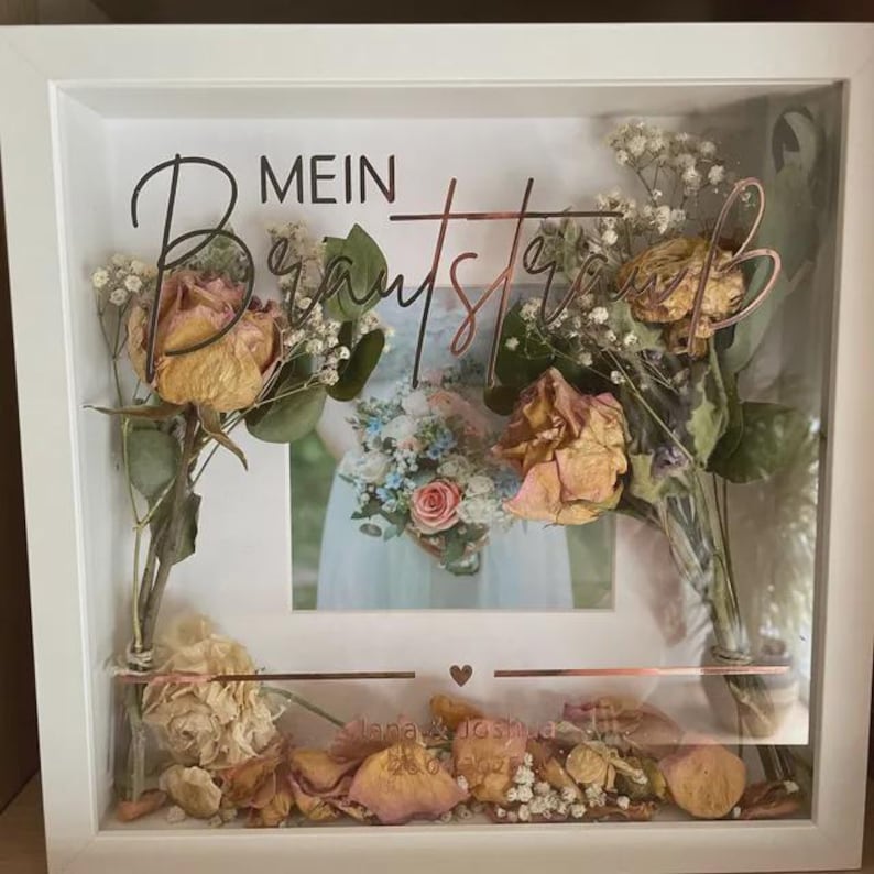 May include: A white framed shadow box with dried flowers and a photo of a bride holding a bouquet. The box includes the text "MEIN Brautstrau&szlig;" in black and copper-colored script. The bottom of the box has the names "Lana & Joshua" and the date "26.06.2021".