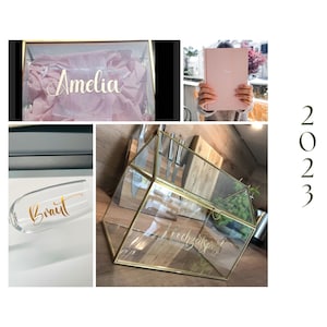 May include: A collection of wedding-themed items. Includes a clear glass box with the name "Amelia" in gold lettering, a pink book, a champagne flute with "Braut" in gold, and a geometric glass terrarium with gold trim.