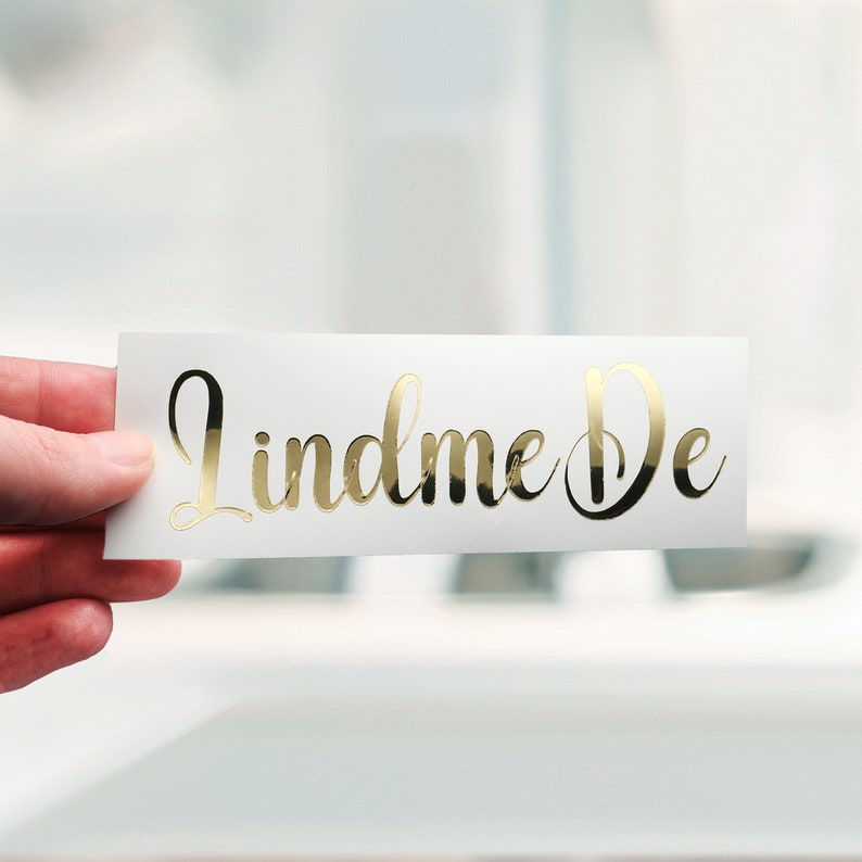 May include: A white rectangular sticker with the gold script text "Lindme De". The sticker is held by a hand, with a blurred background. The gold lettering has a reflective, shiny appearance.