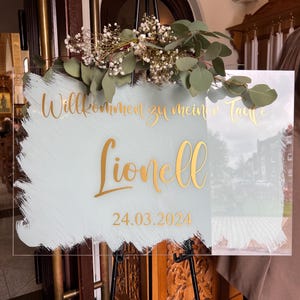 May include: A clear acrylic sign with gold lettering and a light blue and white painted background. The sign reads "Willkommen zu mein Taufe Lionell 24.03.2024". The sign is decorated with greenery and flowers.