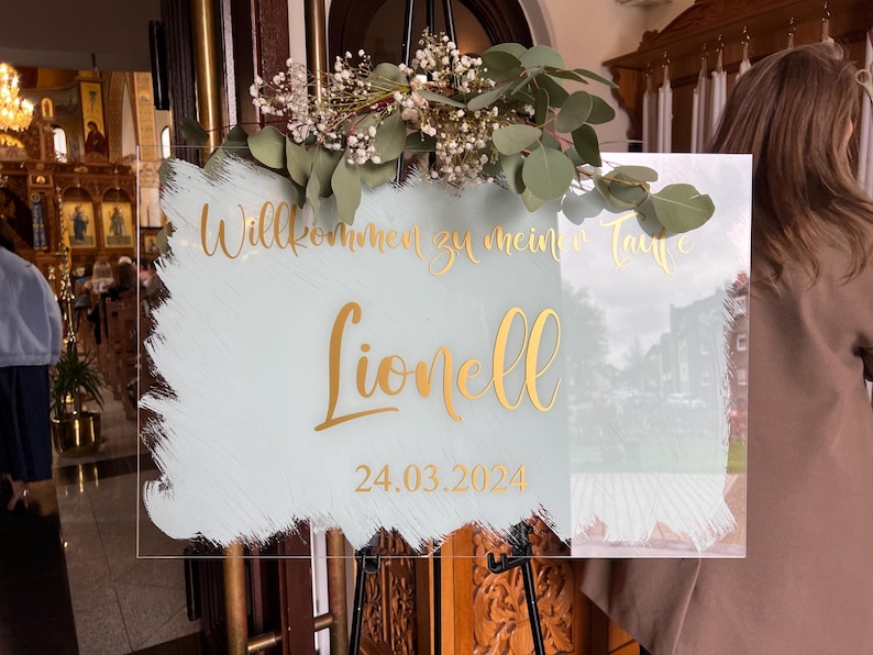 May include: A clear acrylic sign with gold lettering and a light blue and white painted background. The sign reads "Willkommen zu mein Taufe Lionell 24.03.2024". The sign is decorated with greenery and flowers.
