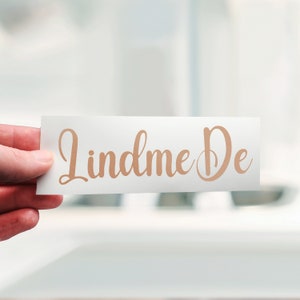 Könnte beinhalten: Eine weiße Karte mit dem Text "Lindme De" in einer kursiv geschriebenen Schrift in hellbrauner Farbe.