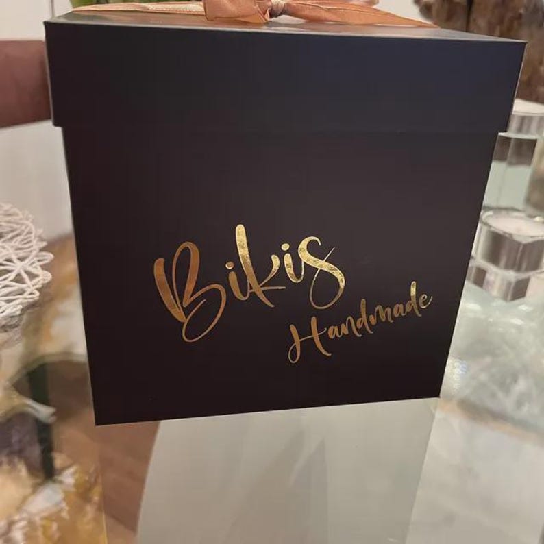 May include: A black square gift box with a gold ribbon and bow. The box has the words "Bikis Handmade" in gold script on the front. The box is sitting on a glass table.
