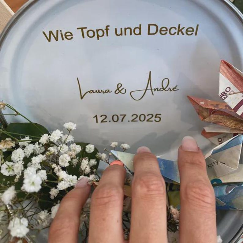 May include: A round, light-colored container with the text "Wie Topf und Deckel", the names "Laura & Andr&eacute;", and the date "12.07.2025". The container is decorated with white flowers and folded paper money.
